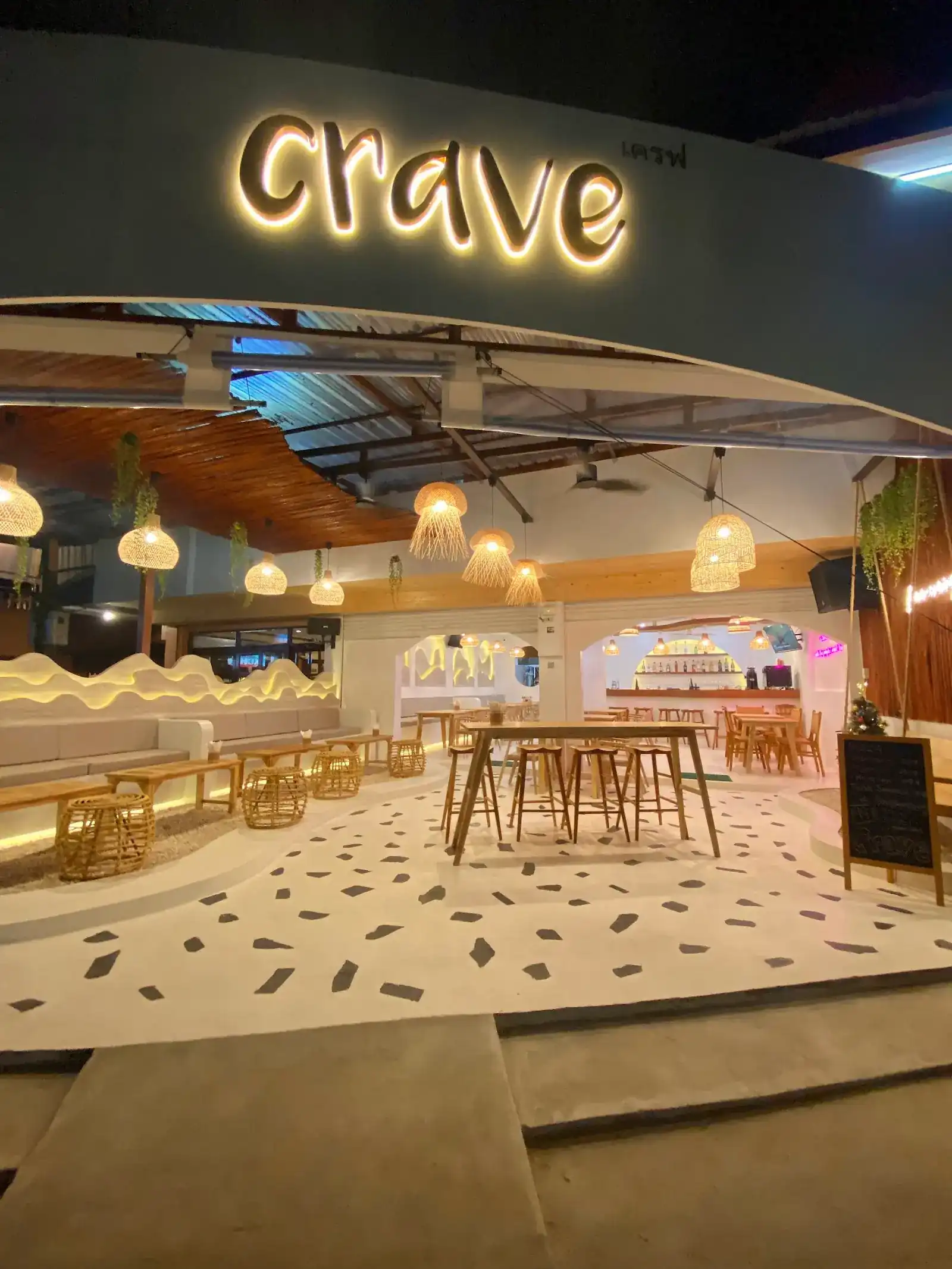 The open front of Crave at night: the lit crave sign above rattan lamps, high tables and a terrazzo floor