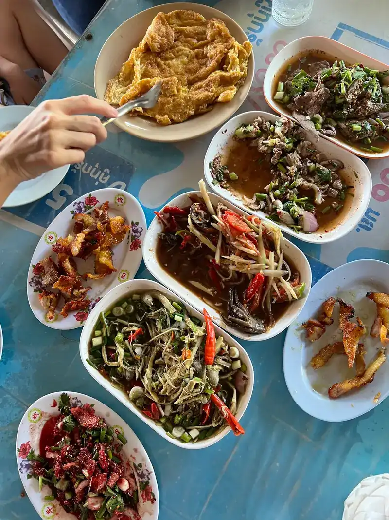 Overhead view of a blue table with an omelette, papaya salad, larb and grilled pork on white plates