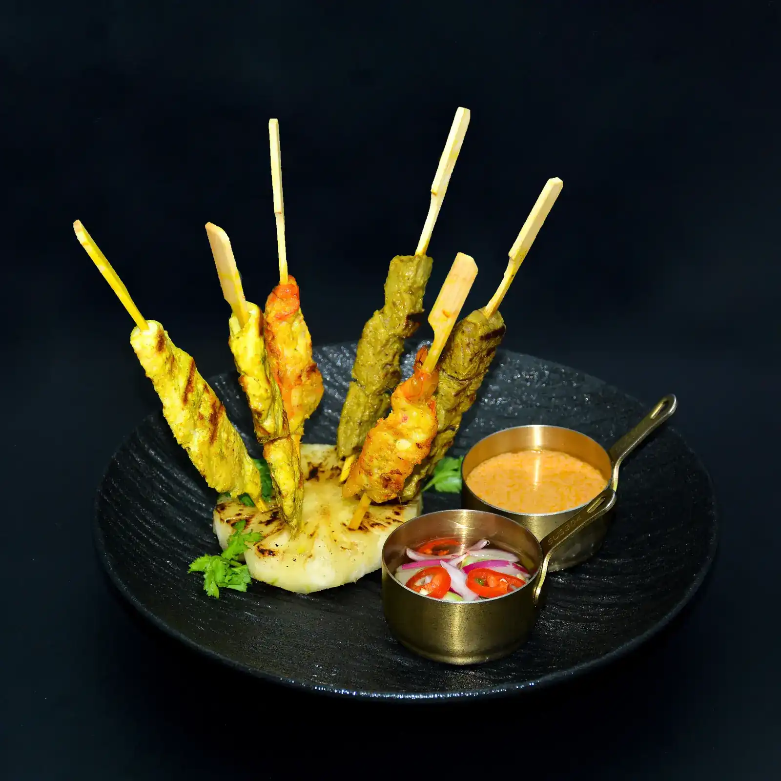 Grilled skewers on a black plate with two small brass pans of sauce