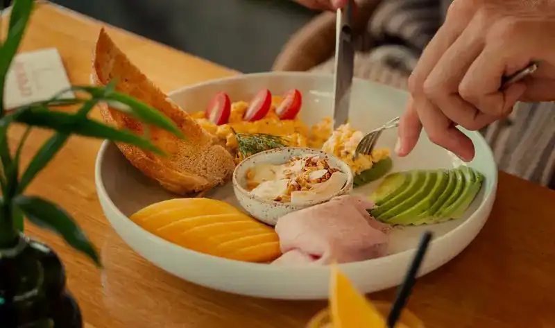 A breakfast plate with scrambled eggs, avocado, ham, mango and sourdough toast