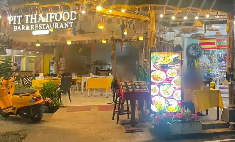 The street front at night: the lit PIT THAIFOOD Bar & Restaurant sign, a yellow scooter and tables in yellow cloths