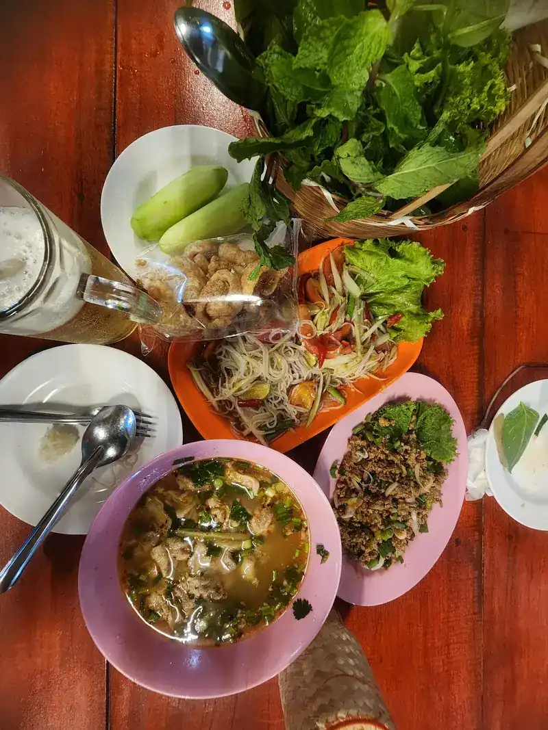 A table seen from above: som tam, larb, a bowl of soup and a basket of fresh greens