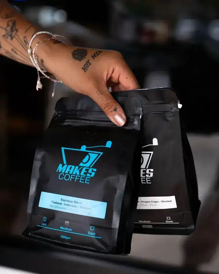 A tattooed hand holding two bags of Makes Coffee espresso blend