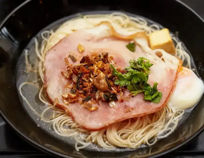 Noodles in clear soup topped with a thick slice of ham, fried shallots, coriander and a soft egg