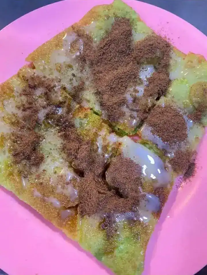 A green roti cut into squares, with condensed milk and chocolate powder on a pink plate