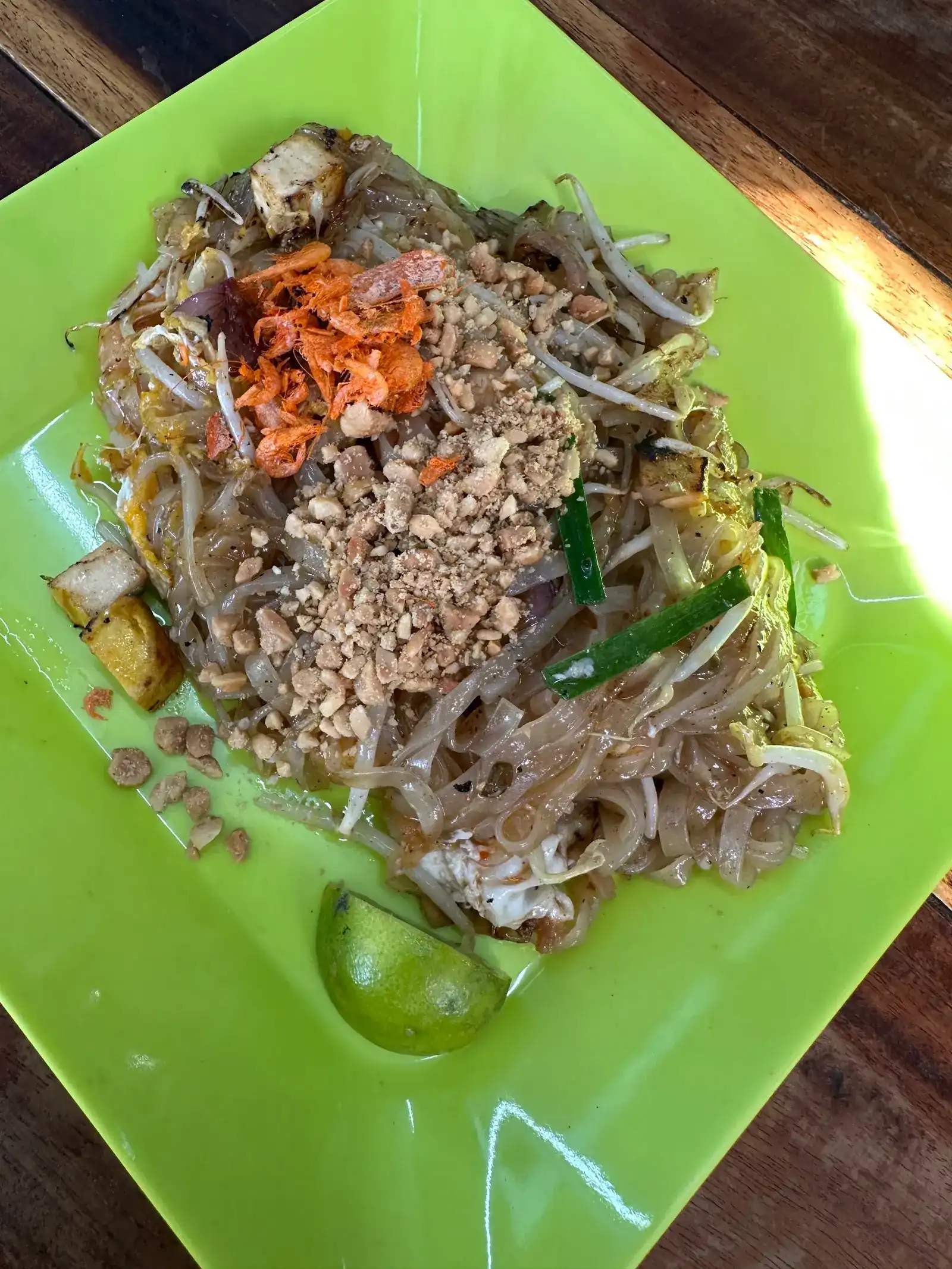 Pad thai on a lime-green square plate, topped with crushed peanuts and dried shrimp, a wedge of lime at the side