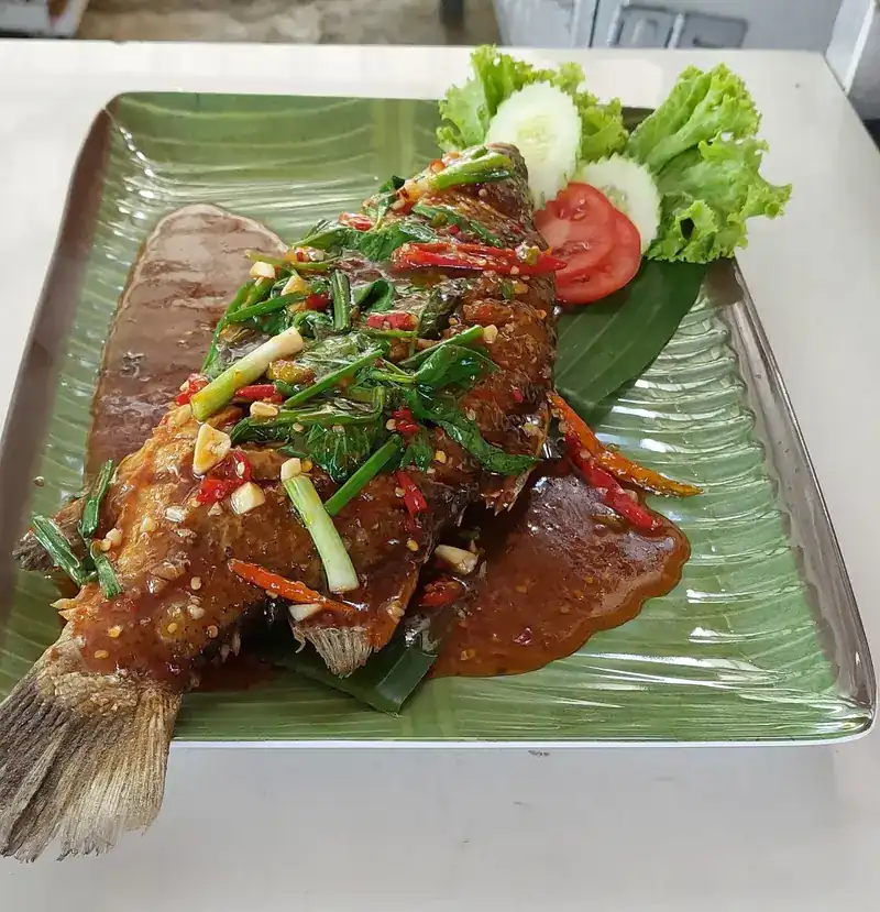A whole fried fish under a thick sauce with chilli, garlic, fresh herbs and spring onion, on a banana-leaf plate
