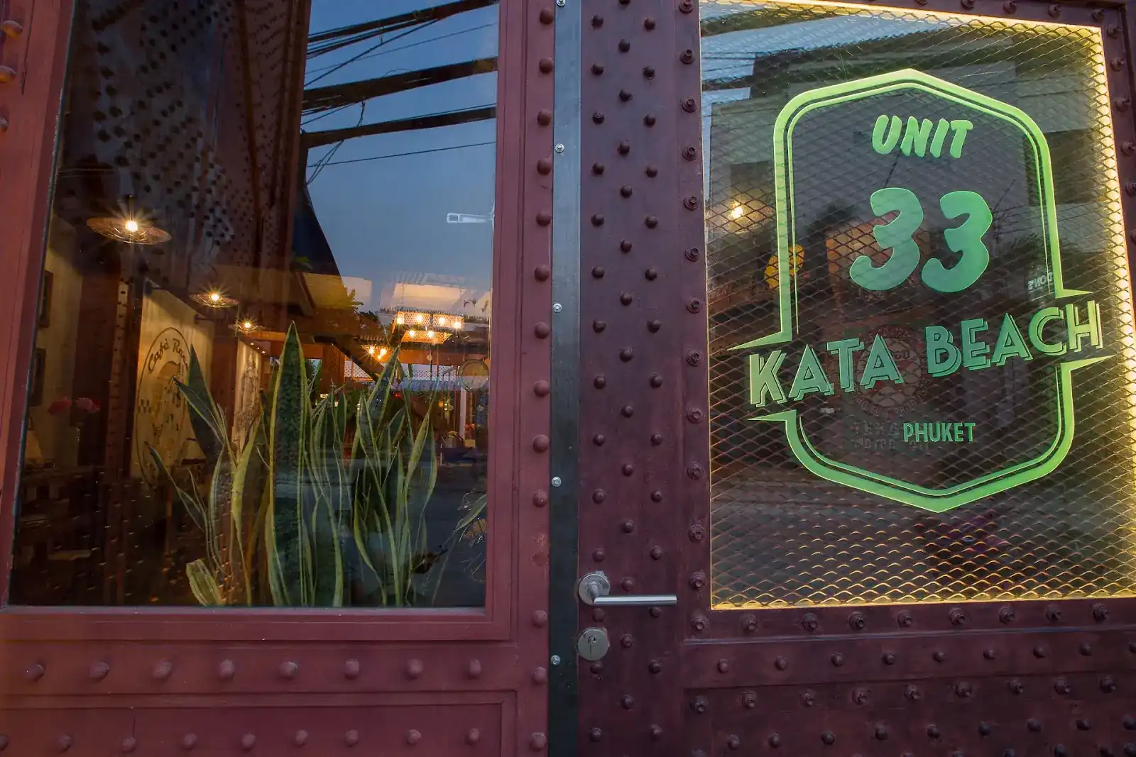 The riveted steel front door of UNIT 33 at dusk, with the green UNIT 33 Kata Beach sign lit in the window