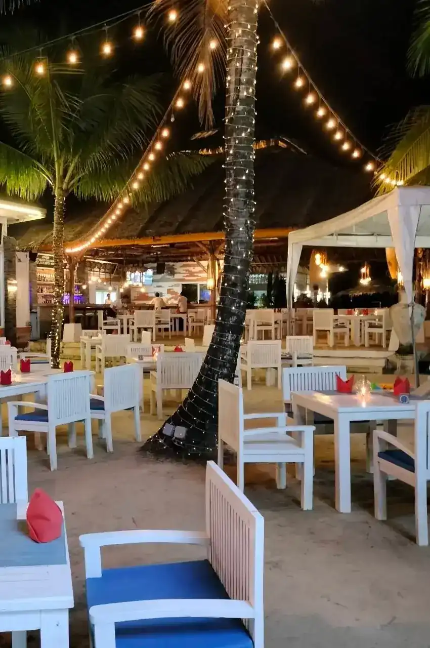 White tables on the sand under palm trees and string lights at night
