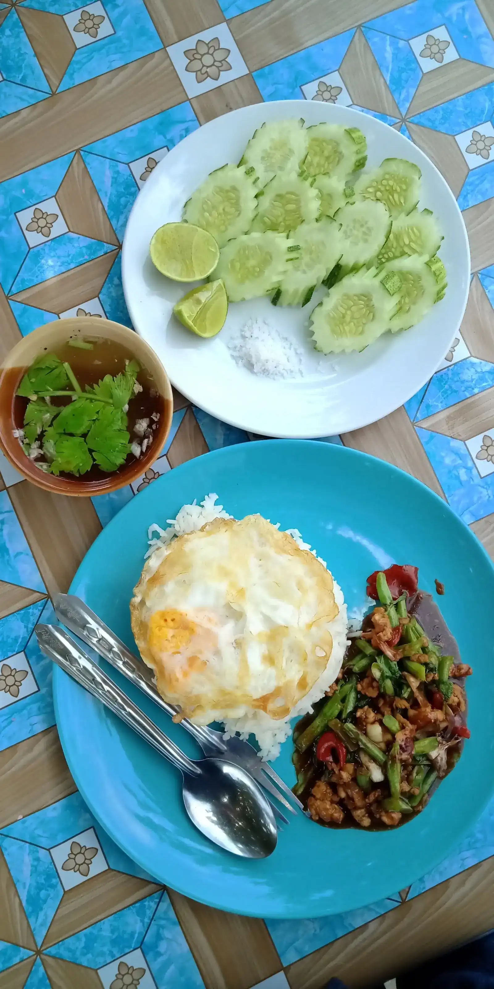 Rice with a fried egg and a stir-fry of minced meat, long beans and chilli, with clear soup, cucumber and lime, on a blue tiled tablecloth
