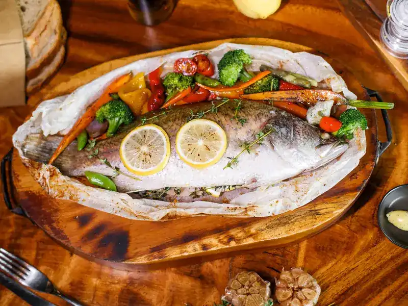 A whole fish baked in paper with lemon, thyme and vegetables, served on a wooden board