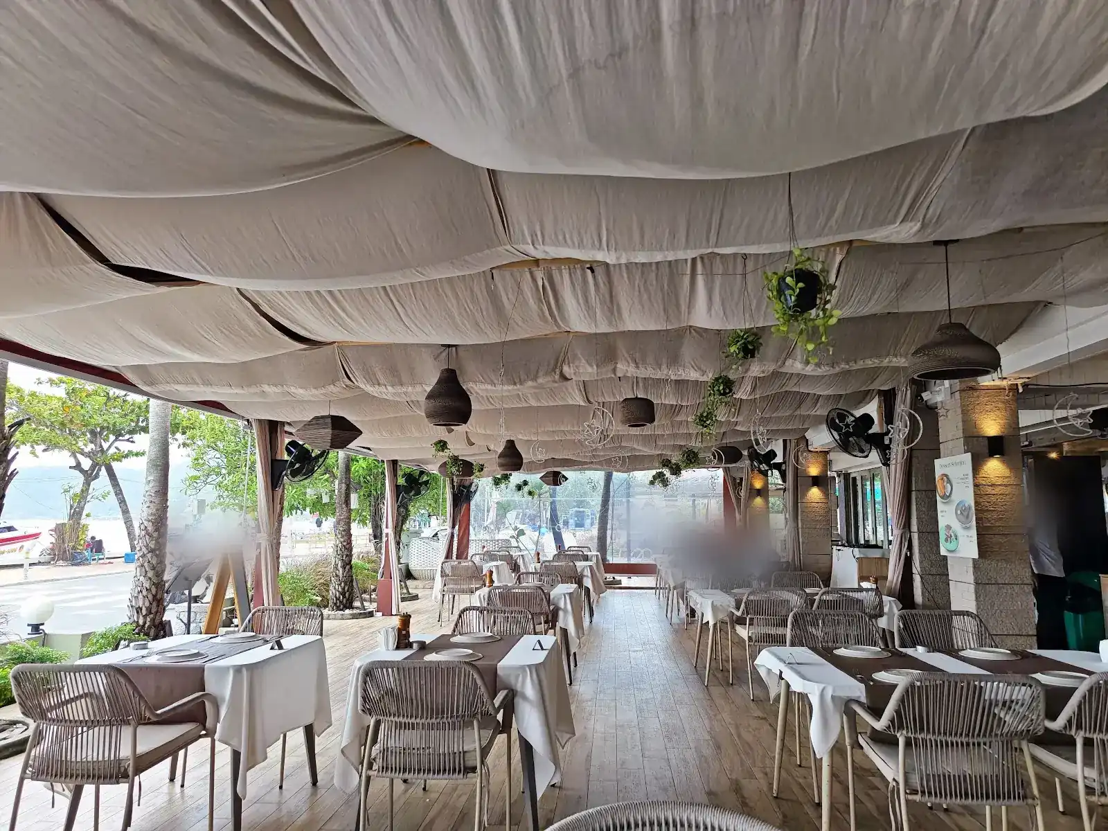 The long terrace by day: a gathered canvas ceiling, pendant lamps and tables laid in white, with palms and the bay across the road