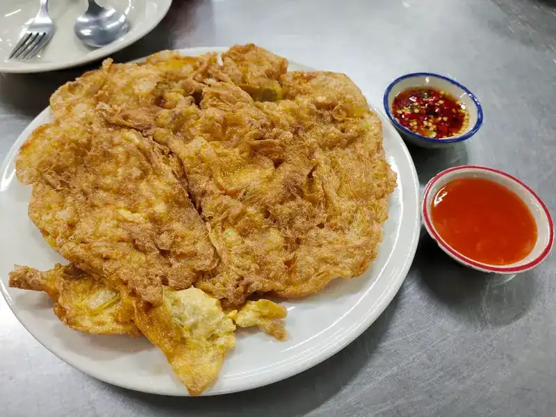 A large golden Thai omelette on a white plate with two small bowls of chilli sauce
