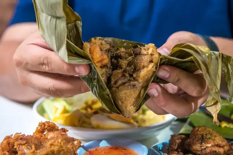 Hands opening a leaf-wrapped ba chang, sticky rice and pork inside