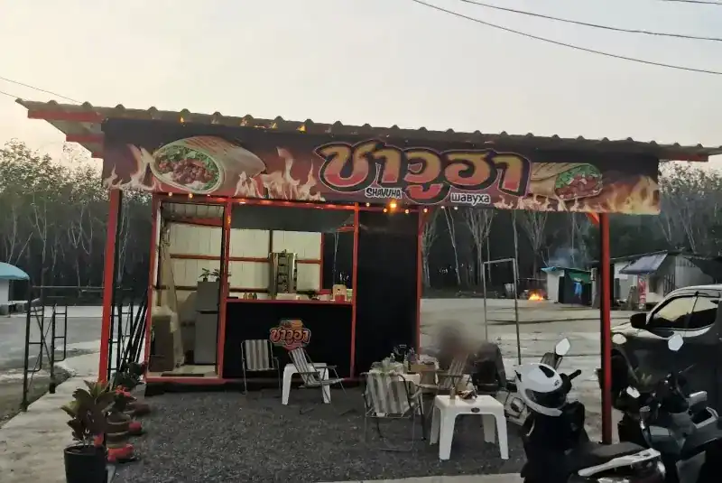 The Shavuha stand at dusk: red steel frame, flame-painted sign, folding chairs and white tables on gravel