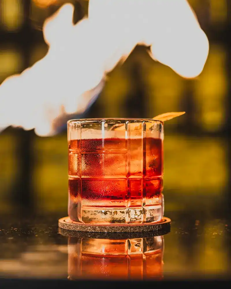 A red cocktail in a short glass on a cork coaster, with a flame above it