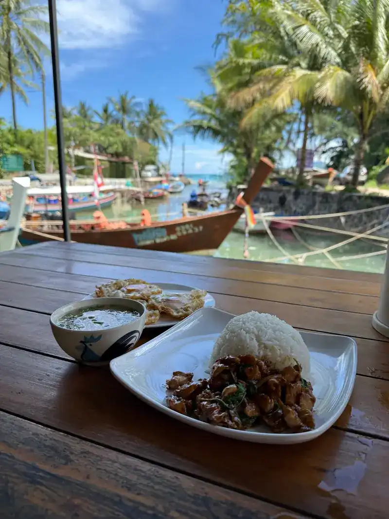 A stir-fry with rice, a bowl of soup and fried eggs on a wooden table, with a longtail boat moored behind
