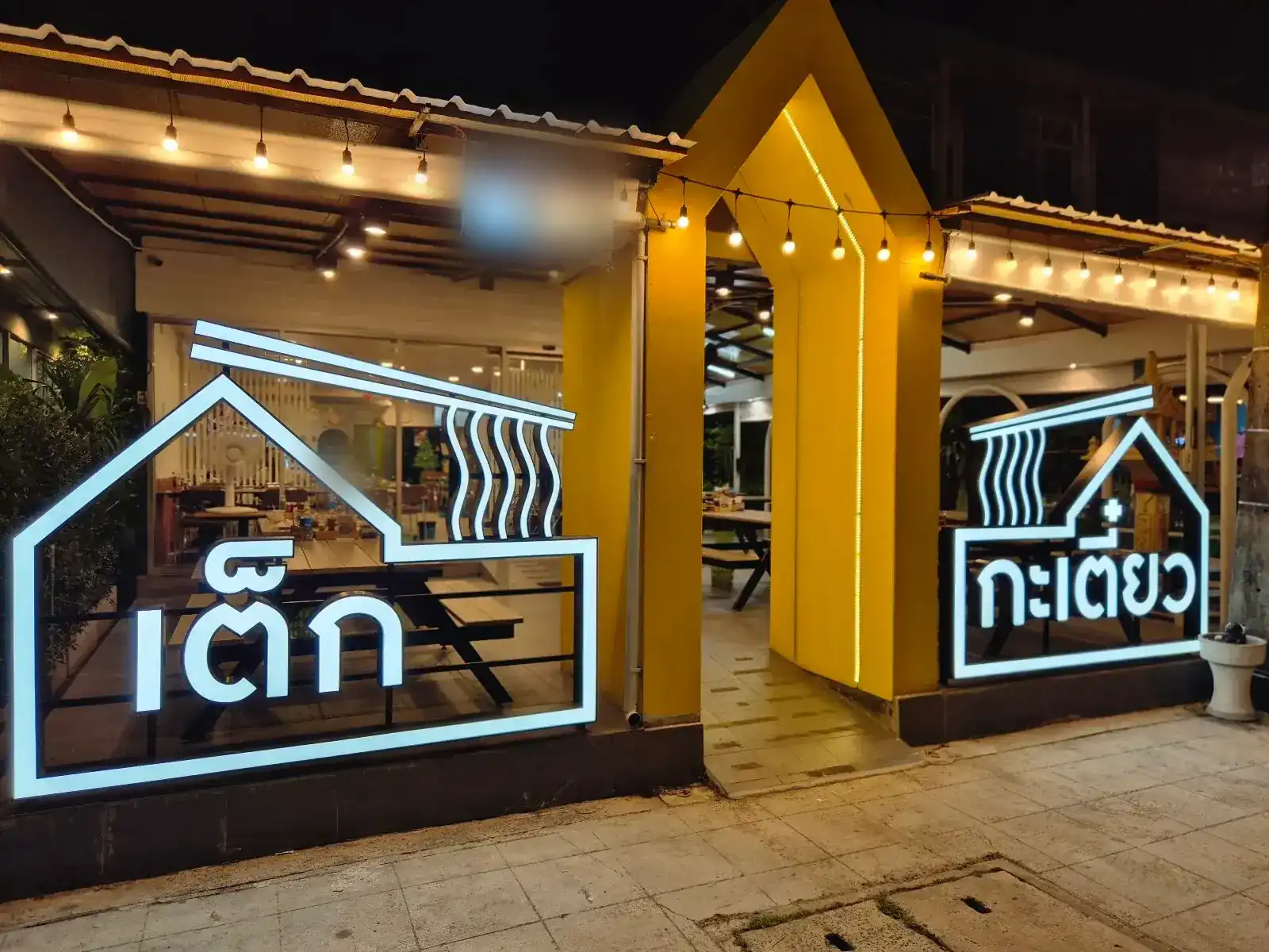 The shopfront at night: two glowing house-shaped signs reading เต็ก and กะเตี๋ยว, with a yellow gabled doorway between them