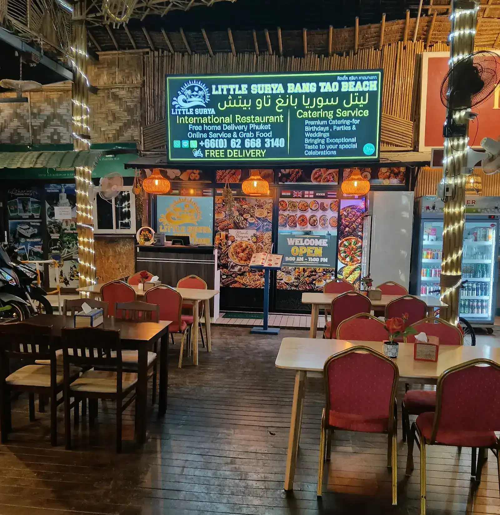 Little Surya's dining room at night: red chairs with gold frames, bamboo walls, woven lamps and the green-lit sign over the counter