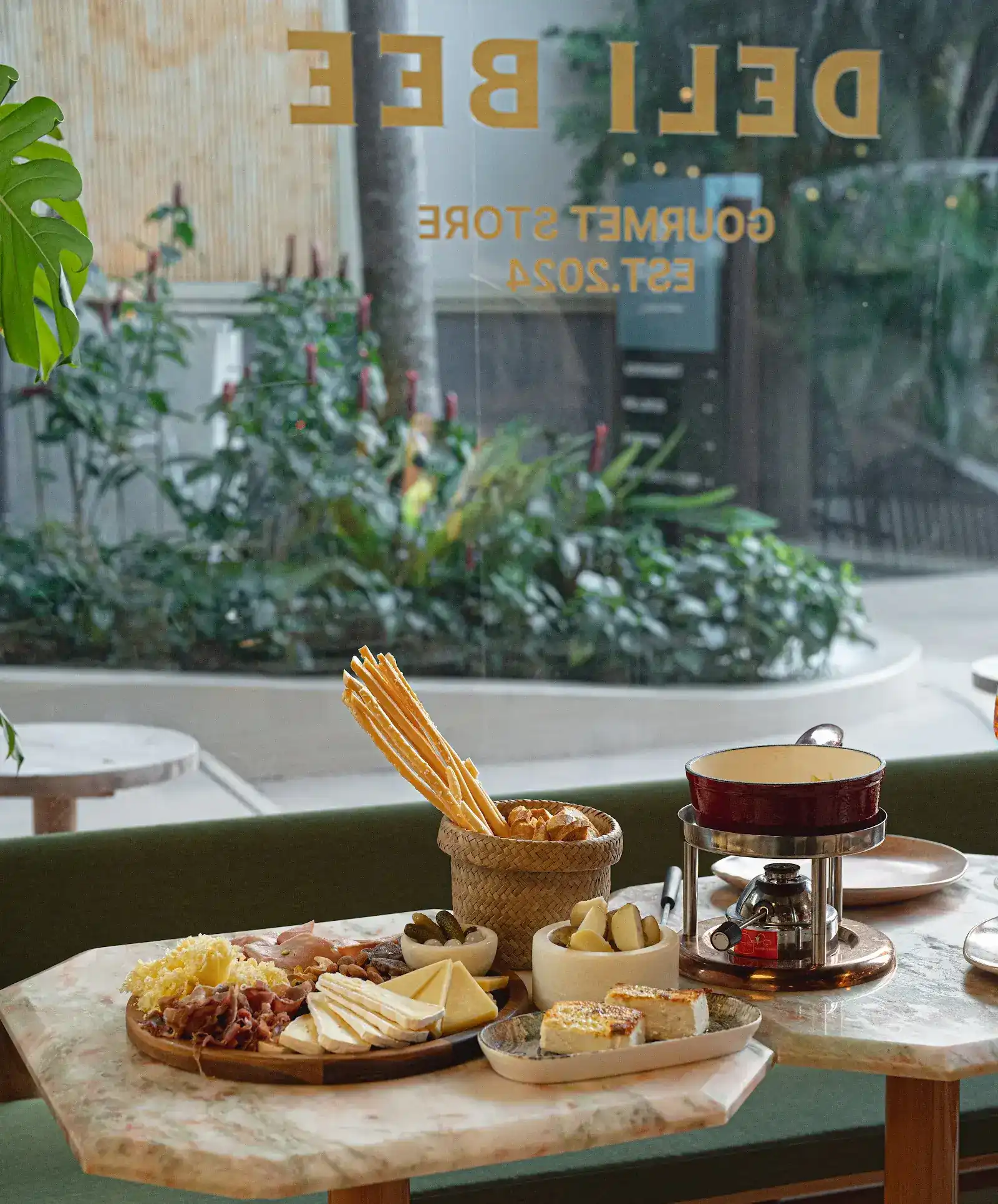 A cheese and cold cut board, grissini and a fondue pot on a pink marble table by the window, with DELI BEE in gold letters on the glass