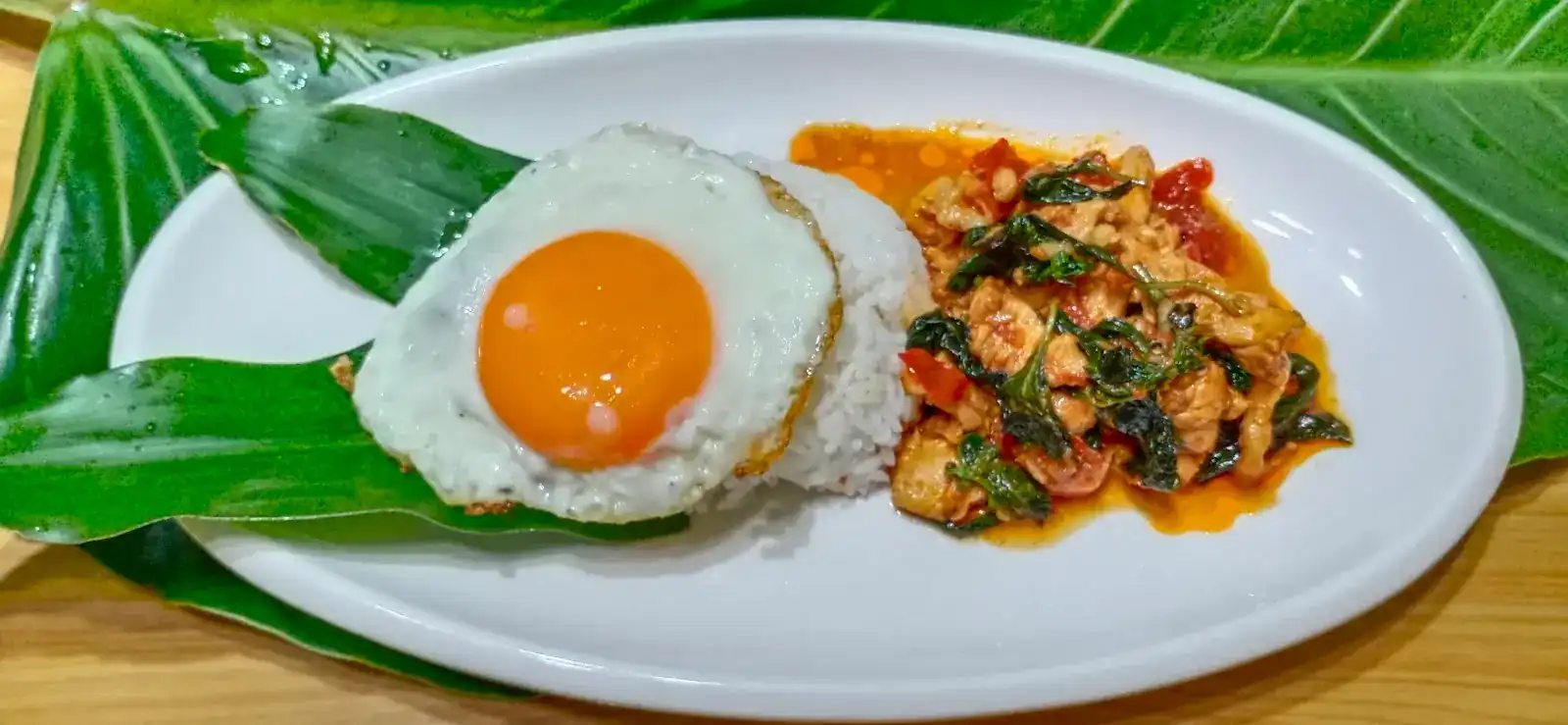 Rice, a fried egg and a spicy stir-fry on a white plate lined with a green leaf