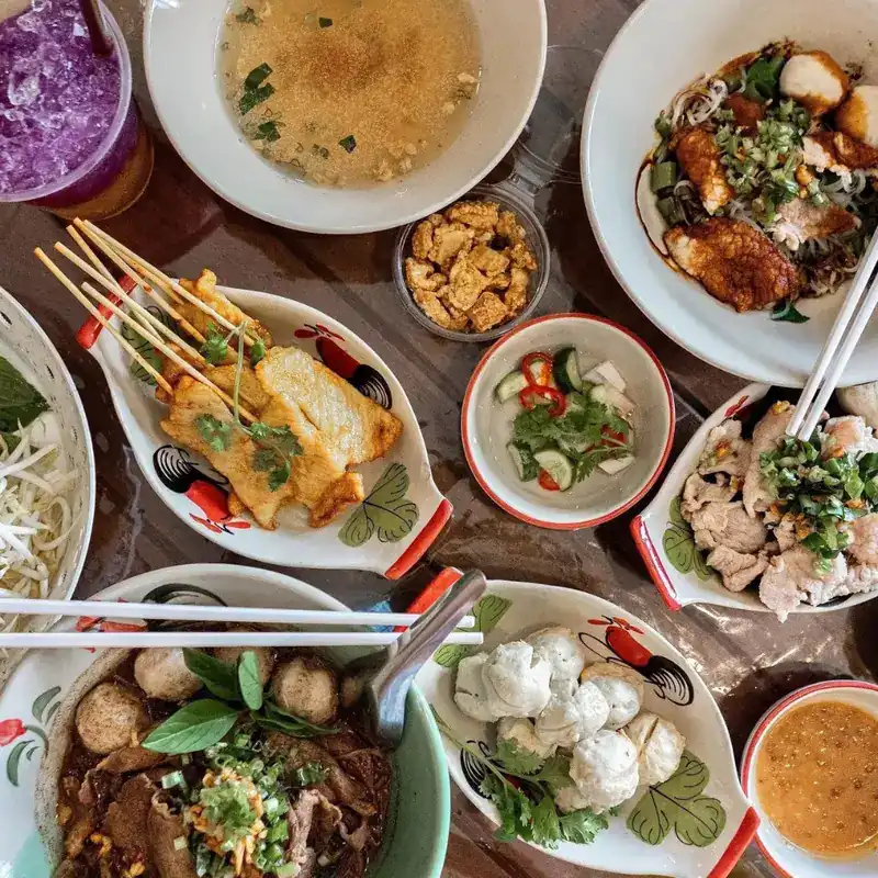 A table of boat noodles, satay, meatballs and side dishes on rooster-pattern plates