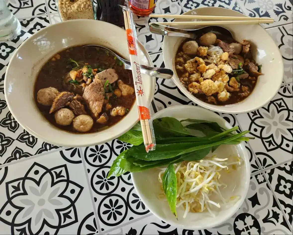 Two bowls of boat noodles with meatballs and crisp pork cracklings, and a plate of fresh herbs and bean sprouts, on a black-and-white tiled table
