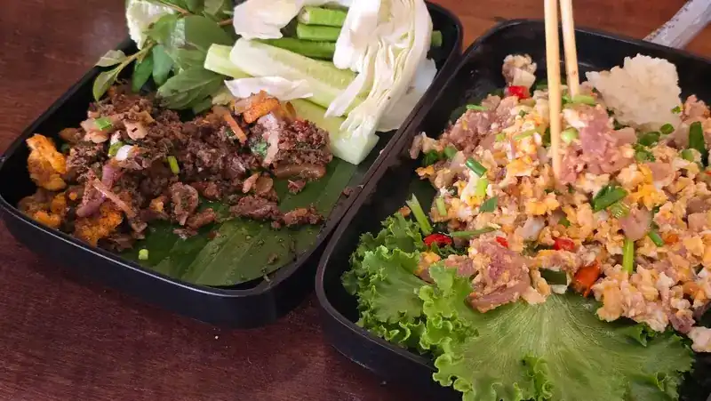 Two trays of minced meat with herbs, one dark and one pale, served on banana leaf and lettuce with cabbage and cucumber