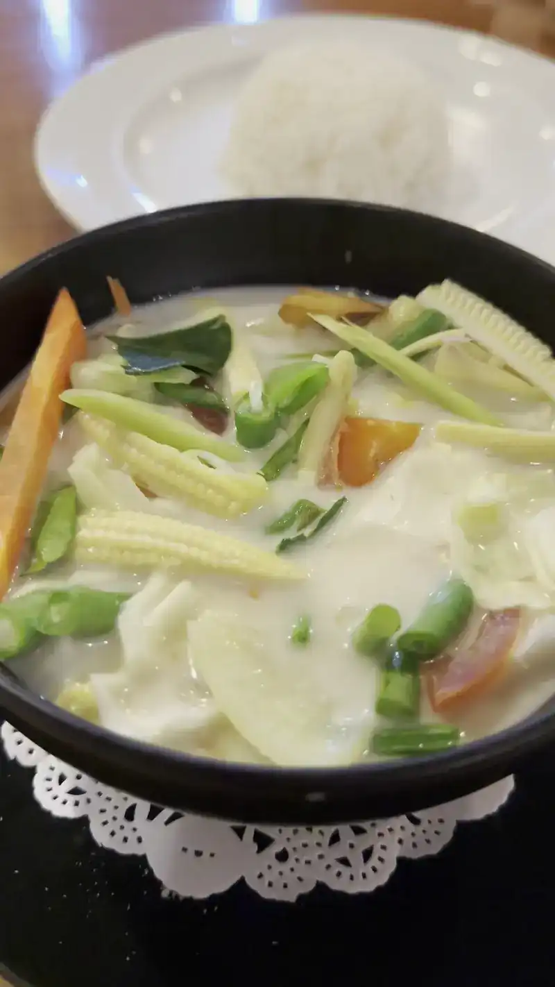 Coconut soup with baby corn, long beans, carrot and tomato in a black bowl, with rice behind