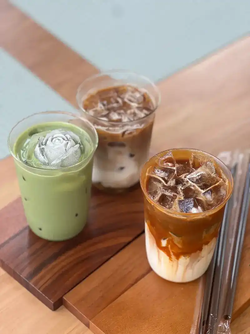 Three iced drinks on a teak board: a matcha latte, a latte and coffee over milk