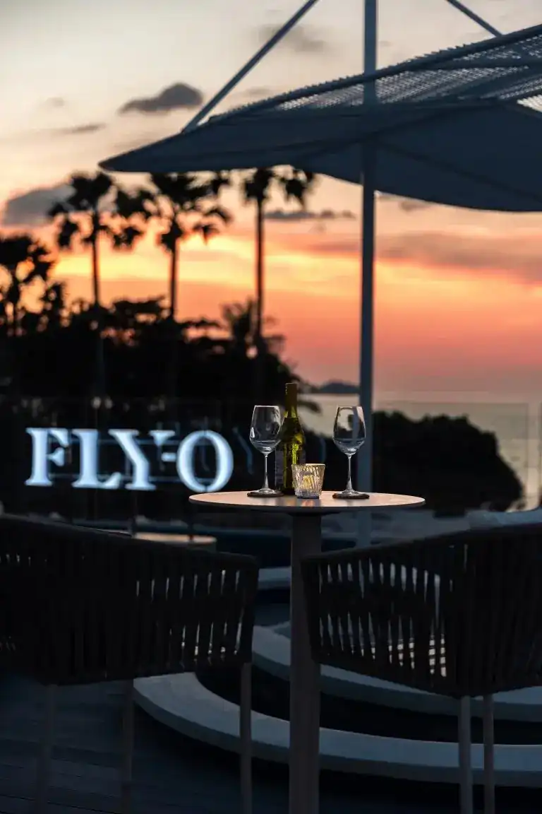 A table for two with wine glasses beside the glowing FLYO sign, palms against a pink sunset over the sea