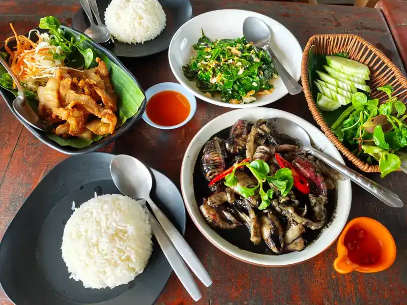 A spread of rice, greens stir-fried with egg, squid in black sauce, a crisp fried dish, fresh vegetables and dips
