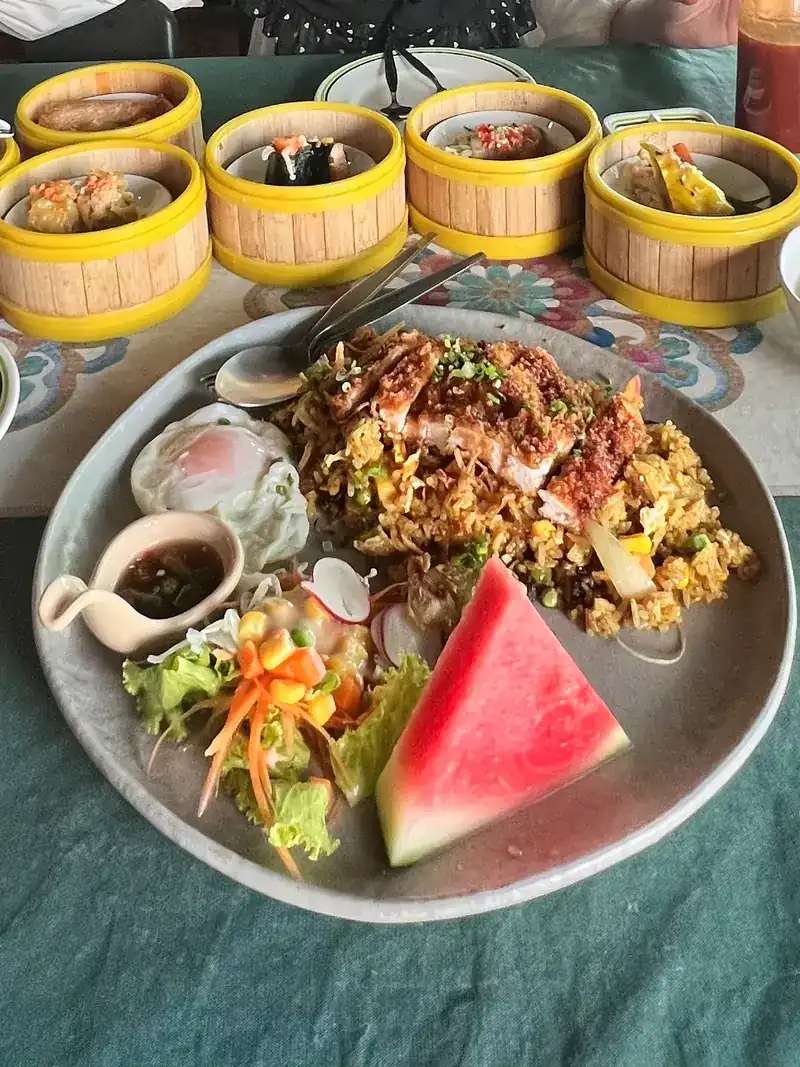 A plate of yellow rice topped with chicken, with salad and watermelon, dim sum steamers behind