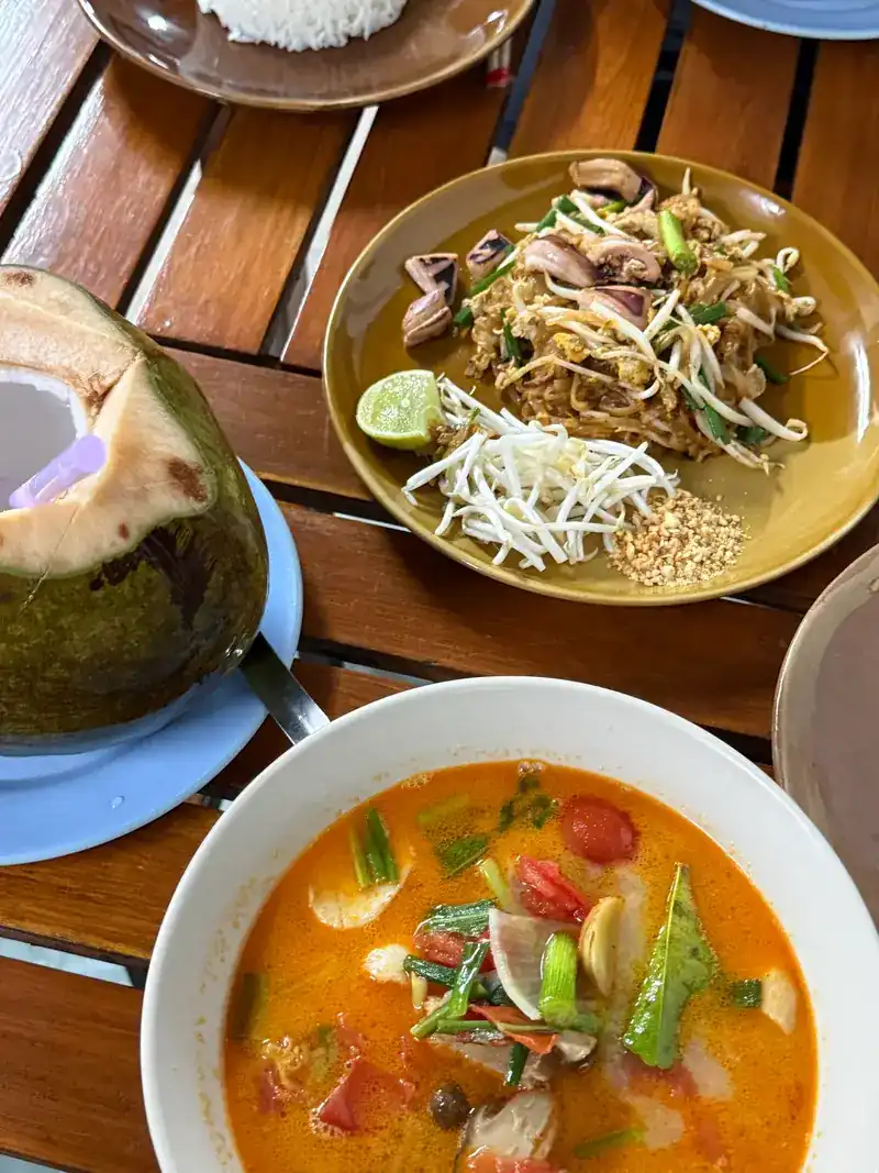 A table with stir-fried noodles, a bowl of red-orange soup with mushrooms, and a fresh coconut