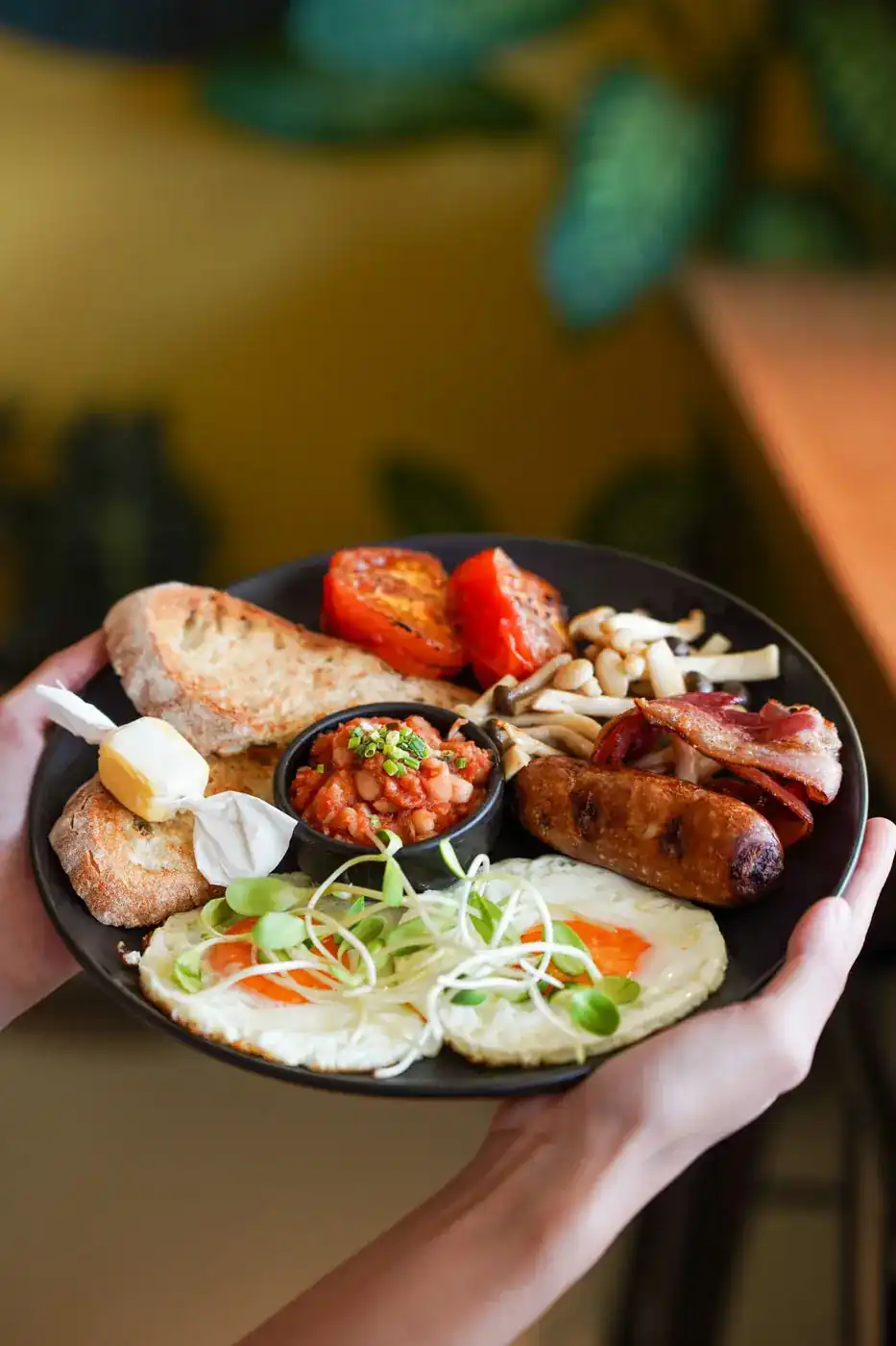 A full breakfast plate held in two hands: toast, fried eggs, sausage, bacon, beans, mushrooms and tomatoes