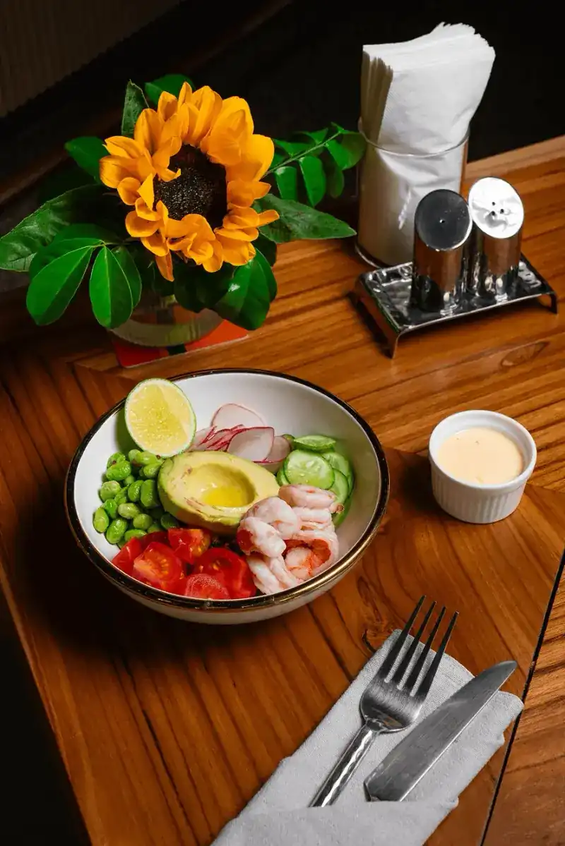 Shrimp poke bowl with avocado, edamame and tomatoes on a wooden table beside a sunflower