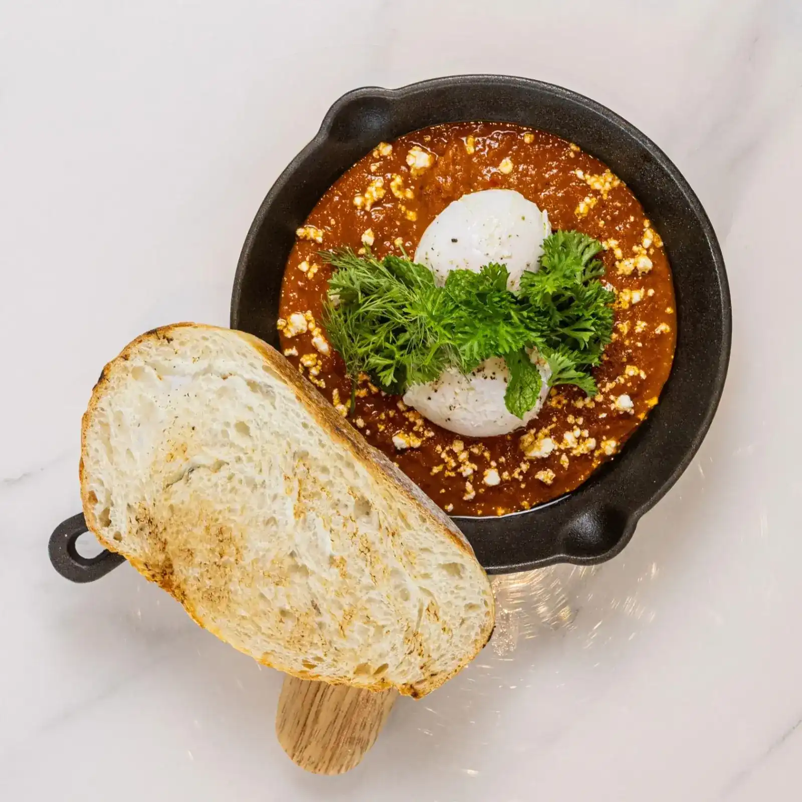 Classic shakshuka in a cast-iron pan with a poached egg, herbs and a slice of toasted bread