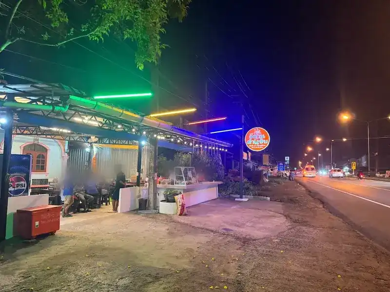 The restaurant at night: an open-air roof hung with lights and coloured neon tubes, a round orange sign, and the road beside it