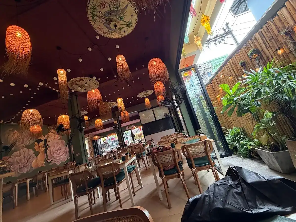 Glowing rattan lanterns hanging from a deep red ceiling over wooden tables, with a bamboo wall and plants at the open side