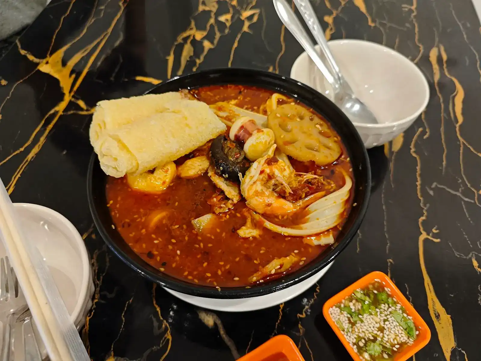 A black bowl of red mala soup with prawn, lotus root, tofu skin and noodles on a black-and-gold table