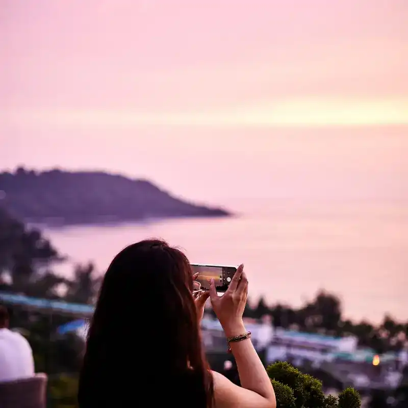 Seen from behind, someone photographs the pink sky over the bay with a phone