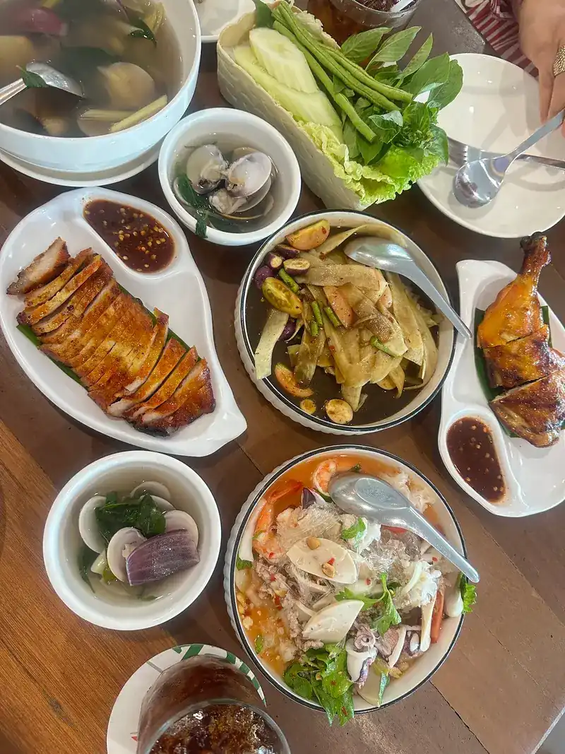 A table of dishes to share: sliced grilled pork, grilled chicken, soups, a seafood salad and fresh vegetables