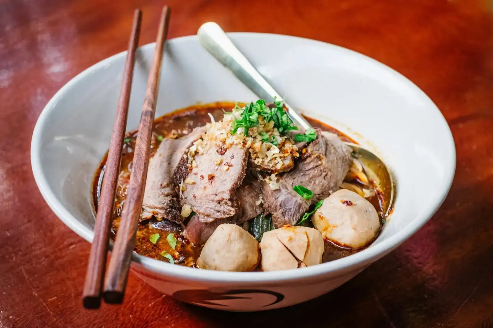 A bowl of boat noodles with slices of beef, beef balls and chopped herbs, chopsticks resting on the rim, on a red wooden table