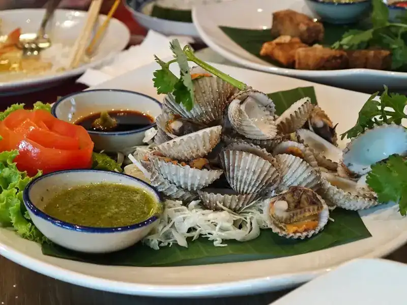 A plate of shellfish on a banana leaf with green seafood sauce and a dark dipping sauce