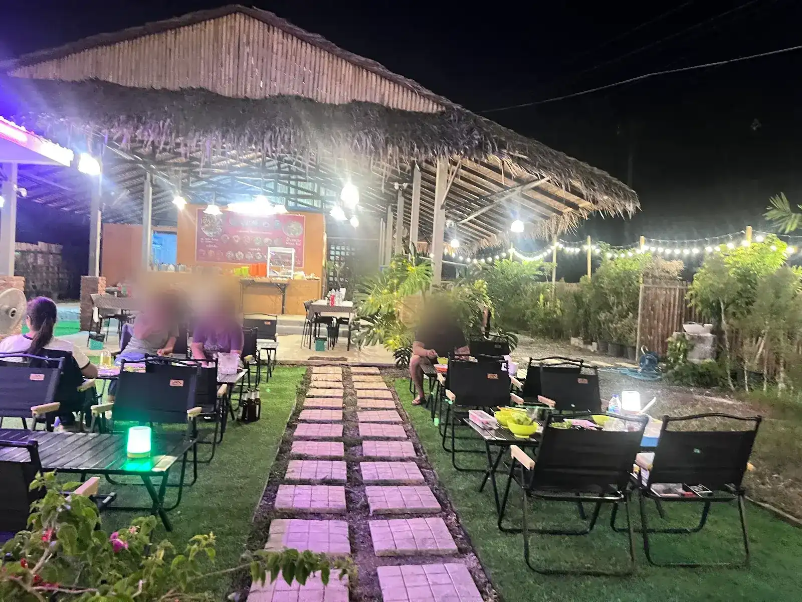 The garden at night: a thatched pavilion lit up, black folding chairs on the lawn and a stone path between the tables