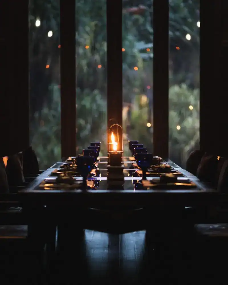A candle lantern on a long dinner table in front of tall windows onto the garden at dusk