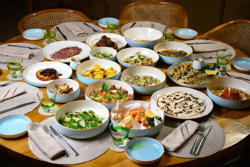 A round table covered with shared plates in sky-blue bowls: pasta, salads, grilled fish and sliced meat