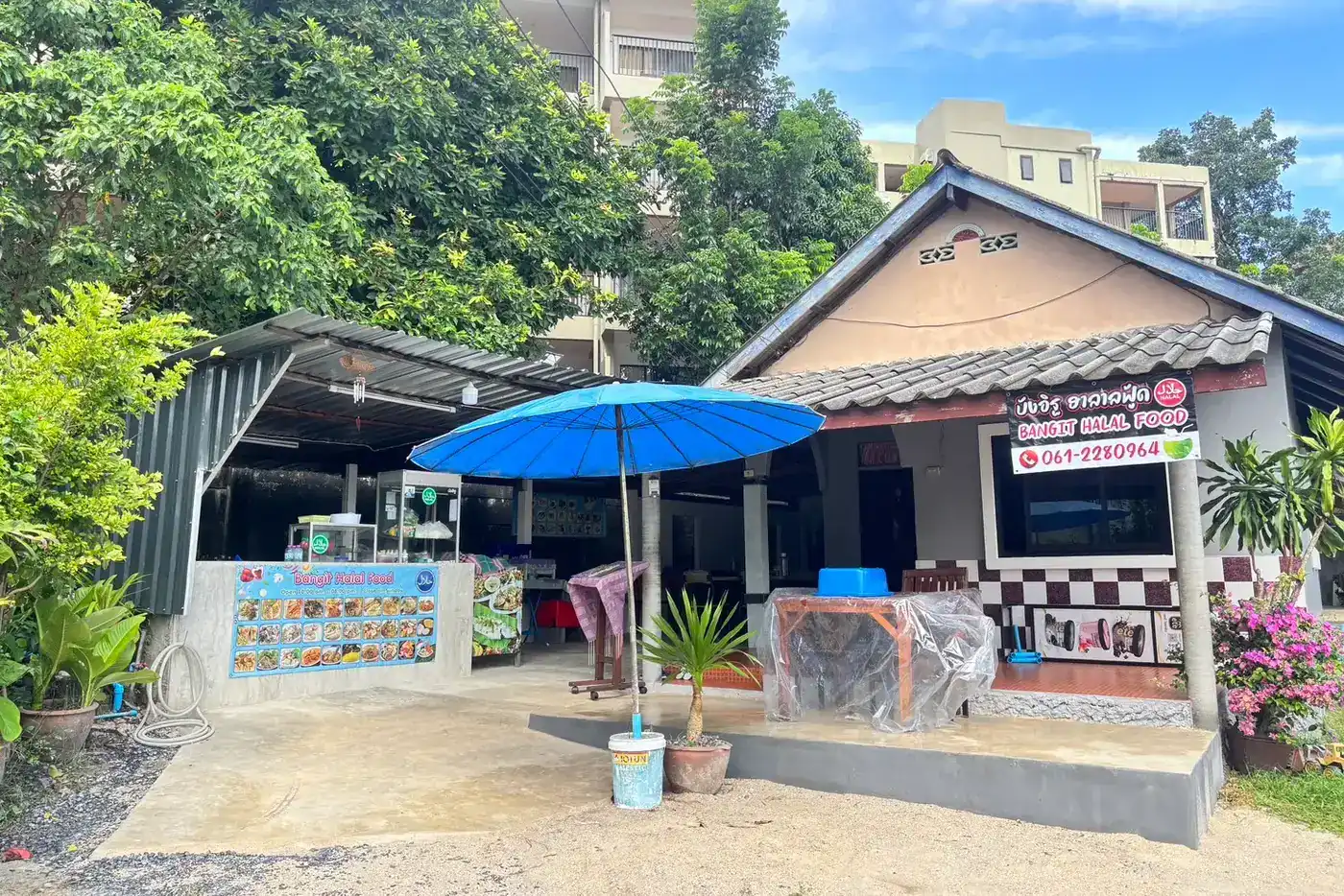 The restaurant from the lane: an open kitchen counter under a metal roof, a big blue umbrella, and the black Bangit Halal Food sign with the phone number