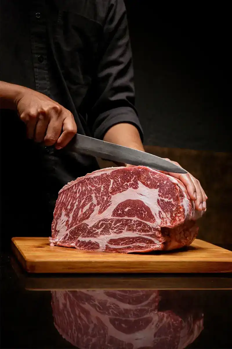 A cook in black slicing a large marbled block of beef on a wooden board