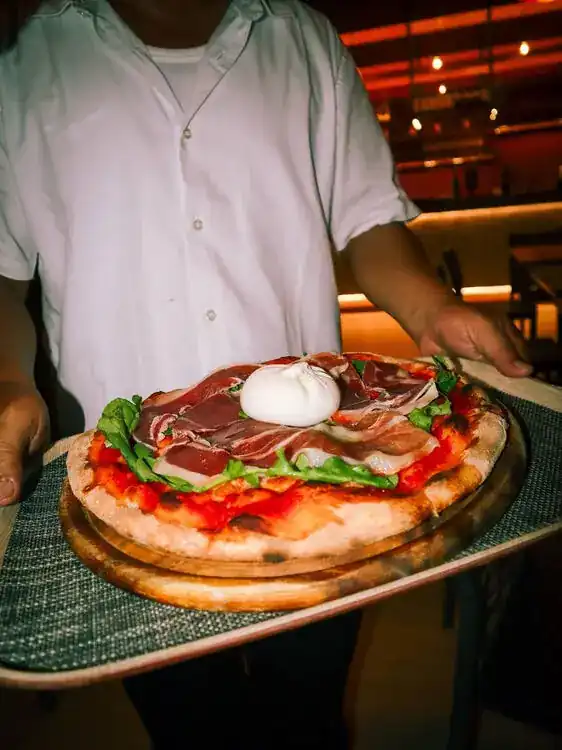 A Roman-style pizza with prosciutto, rocket and burrata carried to the table on a wooden board